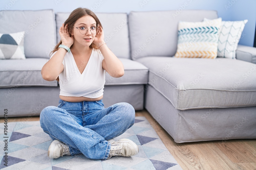 Young caucasian woman sitting on the floor at the living room trying to hear both hands on ear gesture, curious for gossip. hearing problem, deaf Young caucasian woman sitting on the floor at the living room trying to hear both hands on ear gesture, curious for gossip. hearing problem, deaf