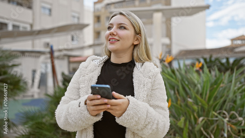 A young blonde woman in a casual outfit looks upwards while holding a smartphone, standing in a green urban park.