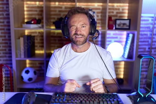 Middle age man with beard playing video games wearing headphones winking looking at the camera with sexy expression, cheerful and happy face.