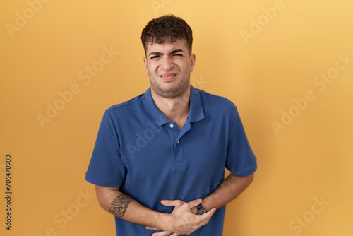 Young hispanic man standing over yellow background with hand on stomach because indigestion, painful illness feeling unwell. ache concept.