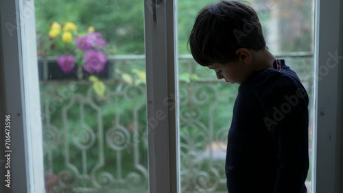 Child stands by window feeling boredom at home looking down with nothing to do © Marco