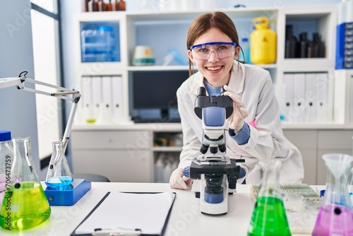Young blonde woman scientist using microscope smiling at laboratory
