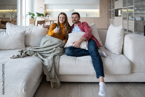 Married couple watch TV together sitting on couch at home enjoying favourite movie series. Pleased woman holding remote control with loving man, ready to change television channel. Family lazy weekend