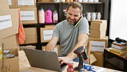 Handsome middle-aged man multitasking on a laptop with a phone at a busy donation center warehouse. photo