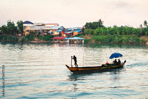 Fishing boat in river in Cambodia with village houses in the background. Mekong near Phnom Penh. Silhouette of fisherman. Khmer culture and tradition. Cambodian life. Cruise, travel and tourism.