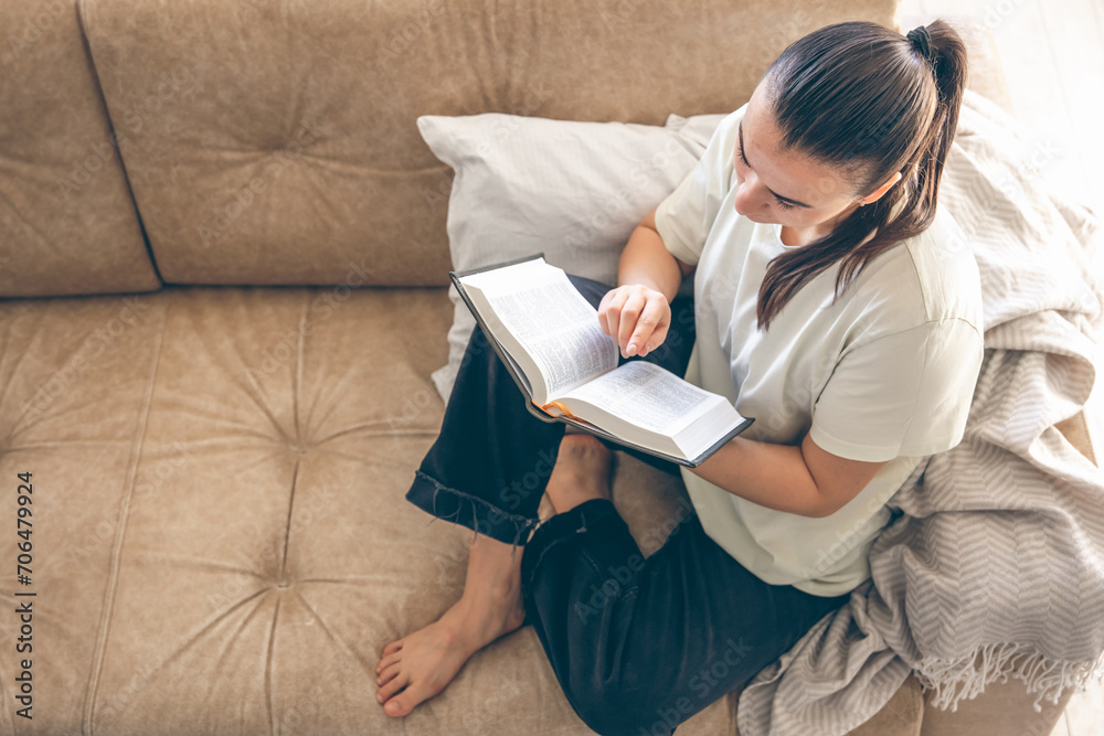 Obraz premium Young Caucasian woman reading a book, sitting on the sofa at home.