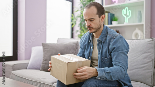 Hispanic bald man with beard holding box sitting on grey sofa in a modern living room © Krakenimages.com