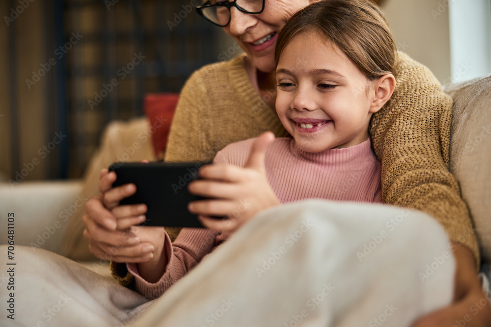 Focus on the smiling little girl, sitting in her grandma's lap, covered in a blanket, using a mobile phone. Focus on the smiling little girl, sitting in her grandma's lap, covered in a blanket, using a mobile phone.