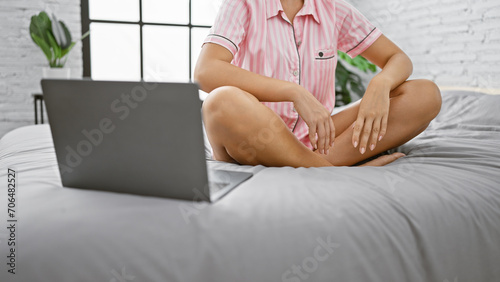 A young blonde woman in casual attire sitting cross-legged on a bed in a well-lit bedroom, with a laptop in front of her. © Krakenimages.com