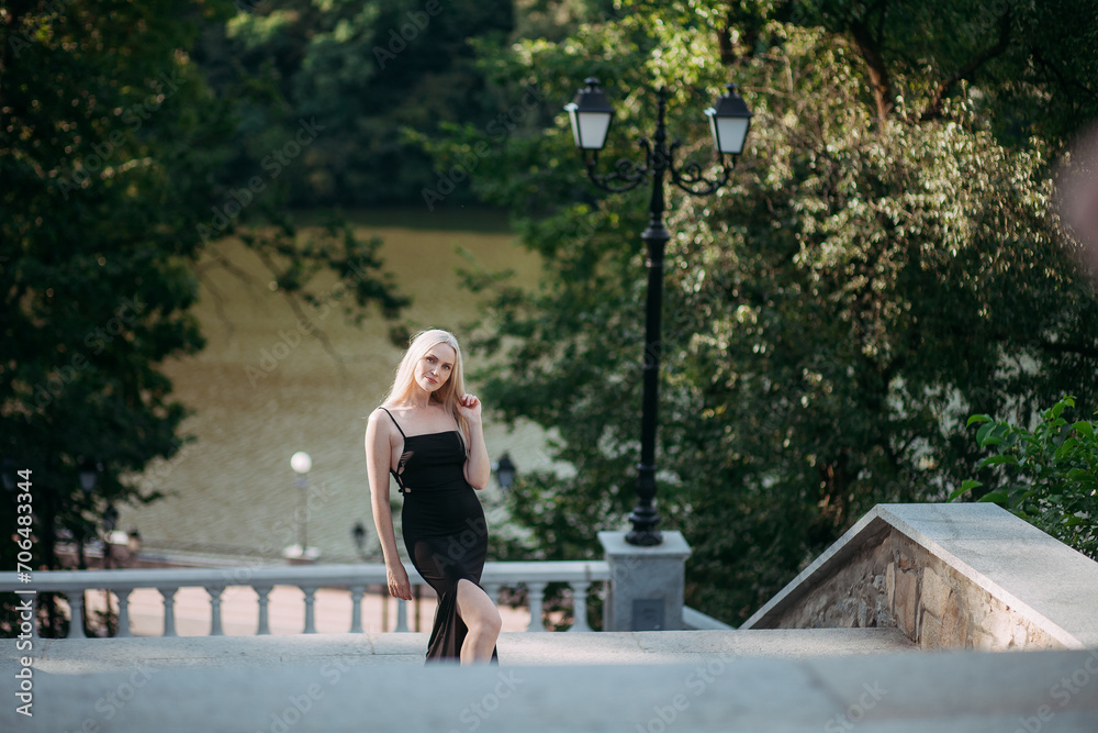 A slender beautiful aged woman with long blond hair, in a black long sexy dress, stands on the stairs in a city park against the backdrop of a picturesque river.
