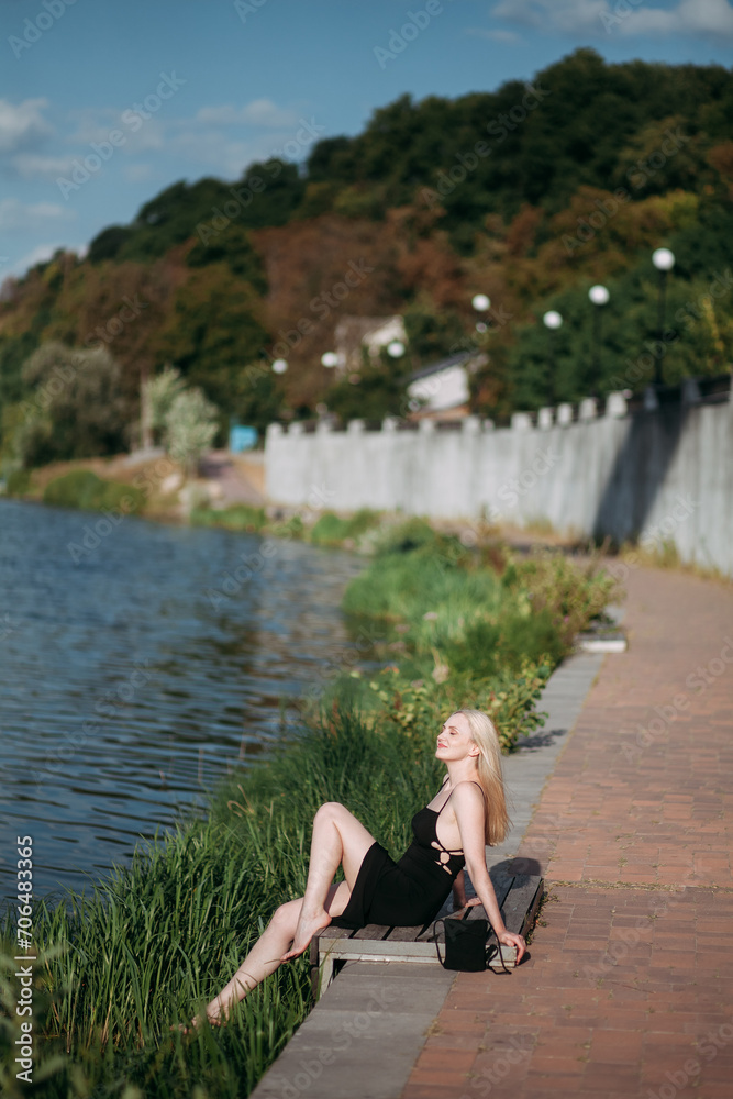 A stylish sexy aged woman with long blond hair and slender long legs, in a black long dress and a black small handbag, sits on the embankment of a picturesque river, on a bright sunny day.