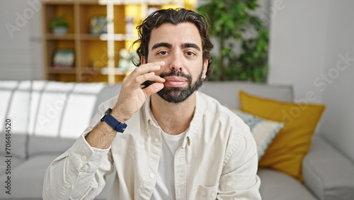 Young hispanic man sitting on sofa doing silent gesture at home
