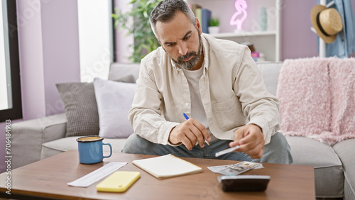 Mature bearded man writing in a notebook in a cozy living room, with coffee and money on the table.