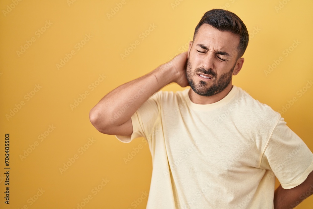 Handsome hispanic man standing over yellow background suffering of neck ache injury, touching neck with hand, muscular pain Handsome hispanic man standing over yellow background suffering of neck ache injury, touching neck with hand, muscular pain