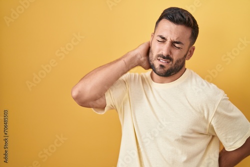 Handsome hispanic man standing over yellow background suffering of neck ache injury, touching neck with hand, muscular pain