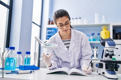 Young beautiful hispanic woman scientist looking sample with loupe writing on notebook at laboratory