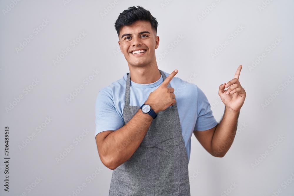 Hispanic young man wearing apron over white background smiling and looking at the camera pointing with two hands and fingers to the side.