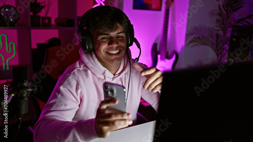 Attractive young hispanic man streamer immersed in digital technology, confidently smiling while having a lively video call on his smartphone in a dark gaming room, joyfully playing games