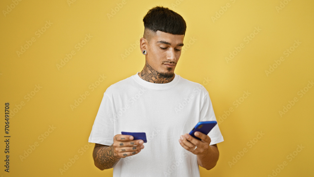 Young, handsome latin man, tattooed and upset, haphazardly uses his credit card and smartphone against isolated yellow wall. Young, handsome latin man, tattooed and upset, haphazardly uses his credit card and smartphone against isolated yellow wall.