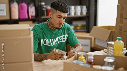 Young handsome tattooed latin man counting donation dollars as a volunteer at a charity center, portraying altruism in his community