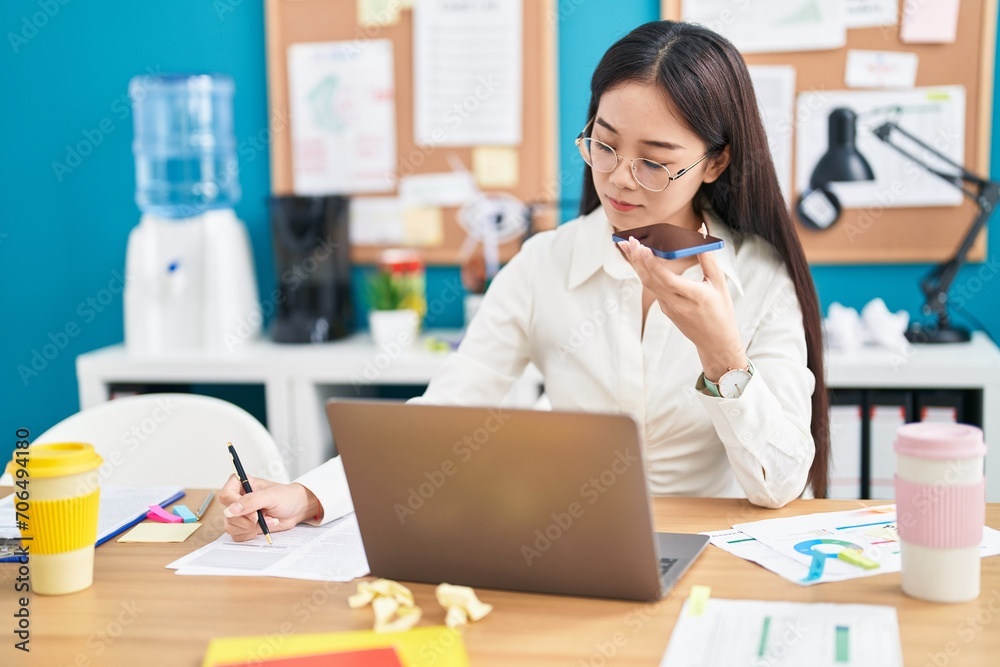 Young chinese woman business worker talking on smartphone writing on document at office