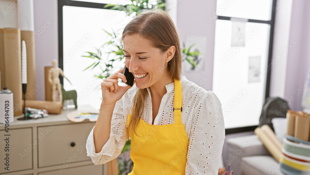 Smiling caucasian woman in a yellow apron talking on a phone indoors, surrounded by artist supplies. Smiling caucasian woman in a yellow apron talking on a phone indoors, surrounded by artist supplies.
