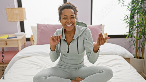 Smiling young adult african american woman with curly hair wearing athleisure sitting cross-legged on a bed in a well-lit bedroom photo