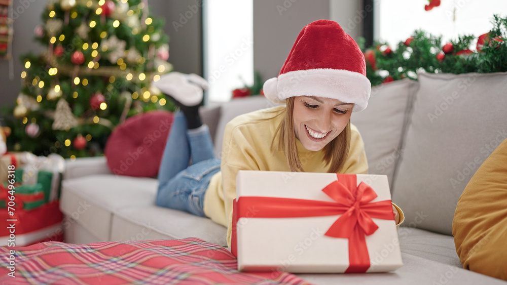 Young blonde woman unpacking gift lying on sofa by christmas tree at home