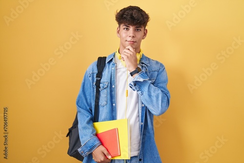 Hispanic teenager wearing student backpack and holding books with hand on chin thinking about question, pensive expression. smiling and thoughtful face. doubt concept. photo