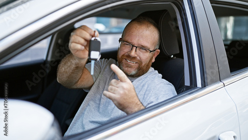 Handsome, bearded, middle-aged caucasian man, smiling confidently, pointing to the key of his new car on a sunny city street- pure enjoyment. © Krakenimages.com