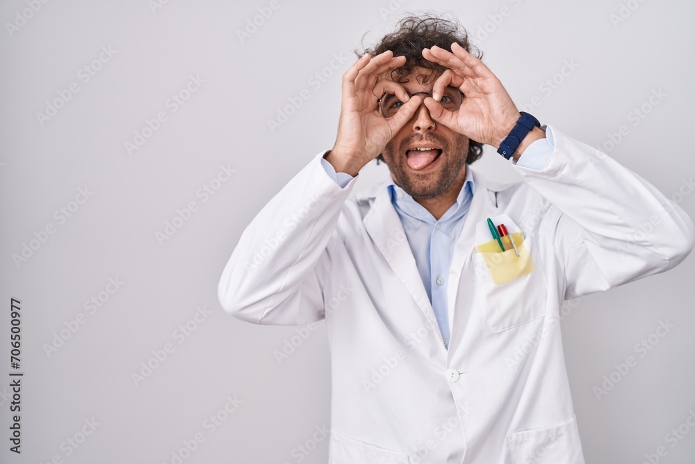 Hispanic young man wearing doctor uniform doing ok gesture like binoculars sticking tongue out, eyes looking through fingers. crazy expression. Hispanic young man wearing doctor uniform doing ok gesture like binoculars sticking tongue out, eyes looking through fingers. crazy expression.