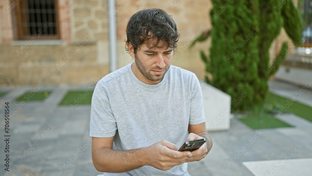 Obraz premium Handsome, young hispanic guy with blond beard gets lost in his smartphone screen while sitting and texting in the lush city park, a portrait of concentrated technology use amidst the green outdoors