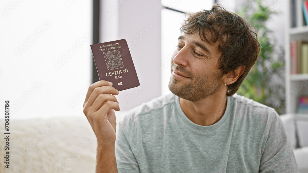 Confident bearded young man sitting on his living room sofa, grinning wide with czech republic passport, overflowing with happiness, ready to embark on an exciting vacation - obrazy, fototapety, plakaty 