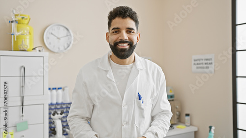 Handsome hispanic man with beard wearing a lab coat standing in a bright hospital room photo