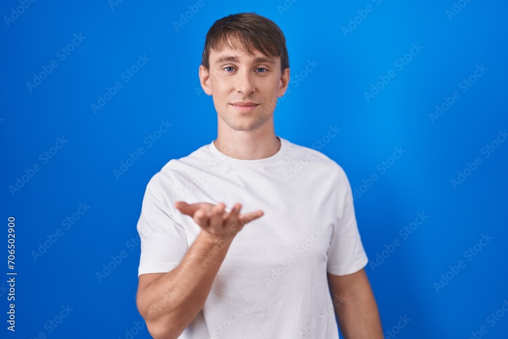 Caucasian blond man standing over blue background smiling cheerful offering palm hand giving assistance and acceptance. Caucasian blond man standing over blue background smiling cheerful offering palm hand giving assistance and acceptance.