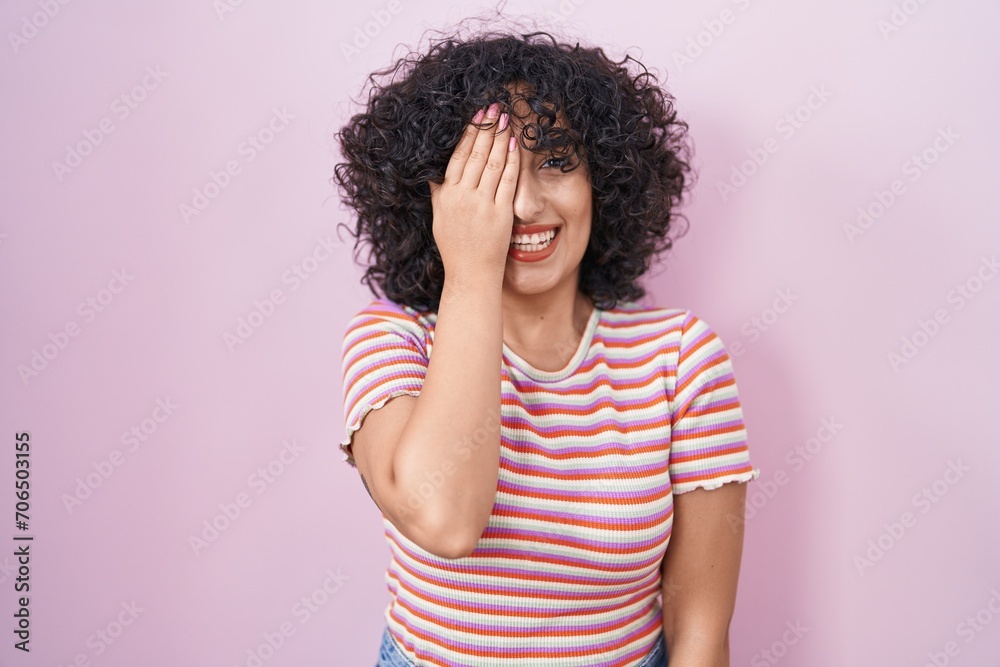 Young middle east woman standing over pink background covering one eye with hand, confident smile on face and surprise emotion. Young middle east woman standing over pink background covering one eye with hand, confident smile on face and surprise emotion.