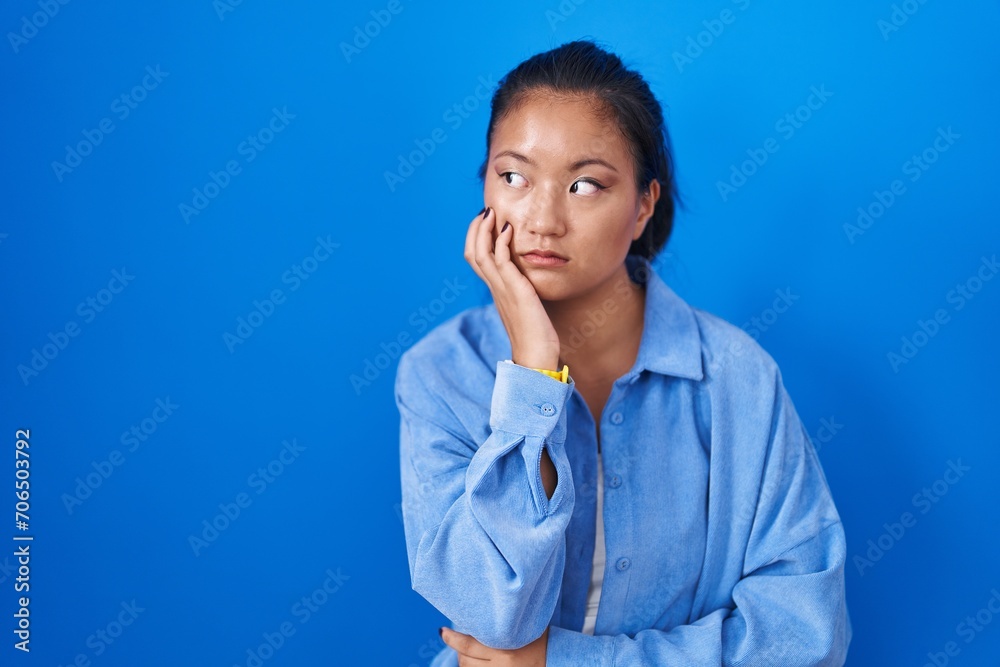 Asian young woman standing over blue background looking stressed and nervous with hands on mouth biting nails. anxiety problem.