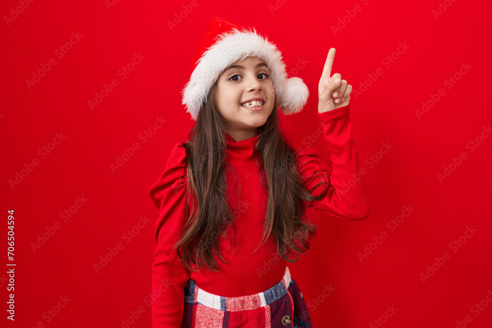 Adorable hispanic girl in christmas hat, gleefully pointing finger up, signaling number one idea success over vibrant red isolated background Adorable hispanic girl in christmas hat, gleefully pointing finger up, signaling number one idea success over vibrant red isolated background