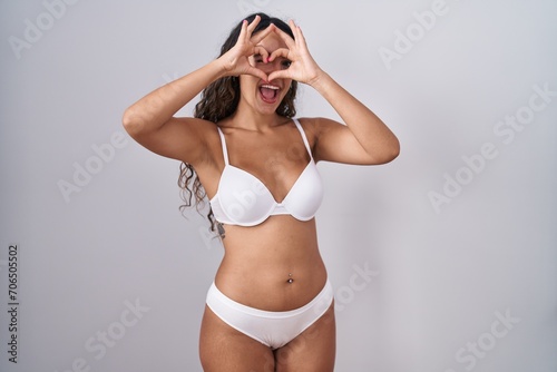Young hispanic woman wearing white lingerie doing heart shape with hand and fingers smiling looking through sign © Krakenimages.com