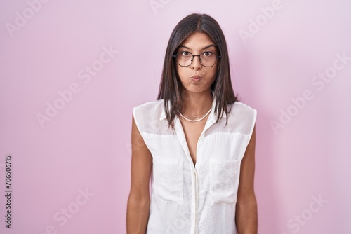 Brunette young woman standing over pink background wearing glasses puffing cheeks with funny face. mouth inflated with air, crazy expression. photo