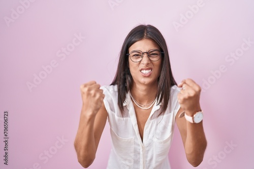 Brunette young woman standing over pink background wearing glasses angry and mad raising fists frustrated and furious while shouting with anger. rage and aggressive concept.