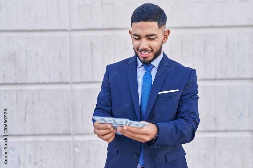 Young latin man business worker smiling confident counting dollars over isolated white background