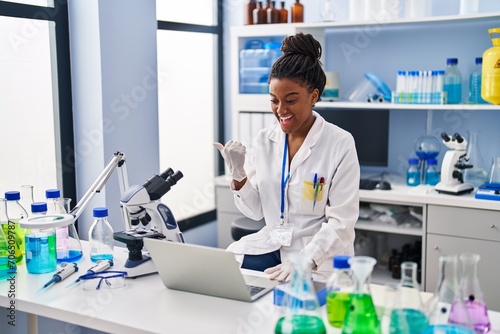 Young african american with braids working at scientist laboratory with laptop pointing thumb up to the side smiling happy with open mouth