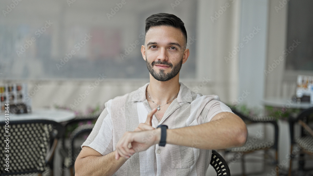 Young hispanic man pointing to watch sitting on table smiling at coffee shop terrace Young hispanic man pointing to watch sitting on table smiling at coffee shop terrace