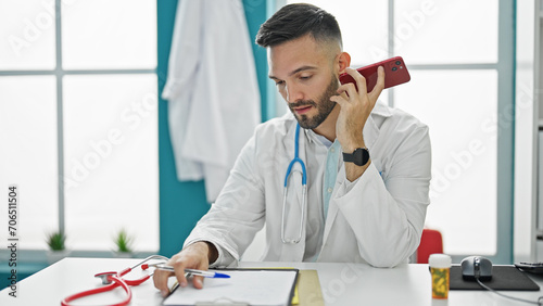 Young hispanic man doctor listening to voice message with smartphone at the clinic