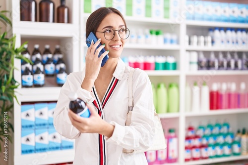 Young beautiful hispanic woman customer talking on smartphone holding medicine bottle at pharmacy