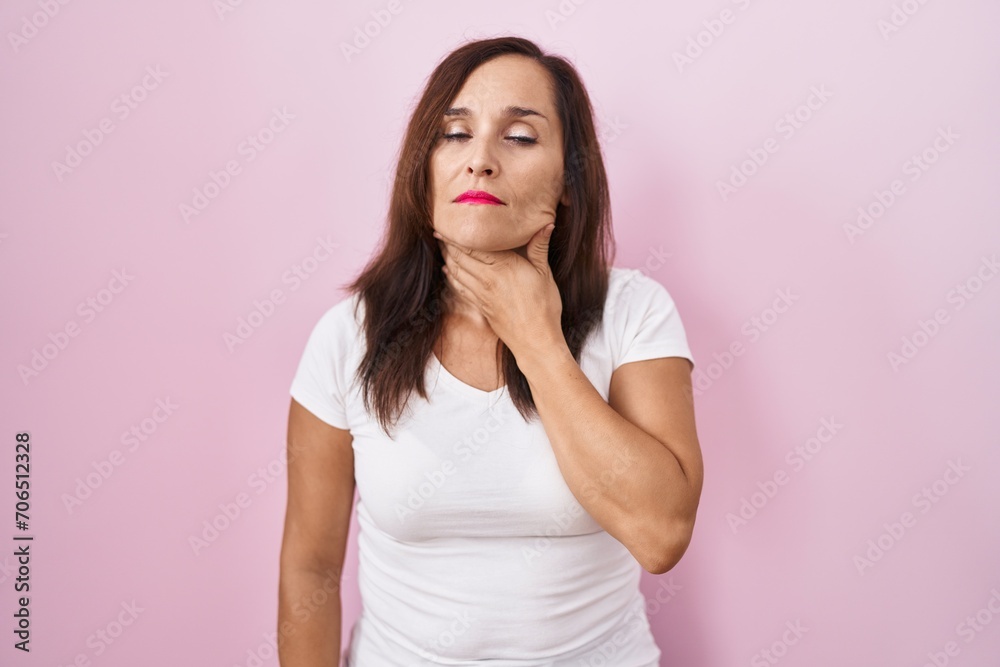 Middle age brunette woman standing over pink background touching painful neck, sore throat for flu, clod and infection Middle age brunette woman standing over pink background touching painful neck, sore throat for flu, clod and infection