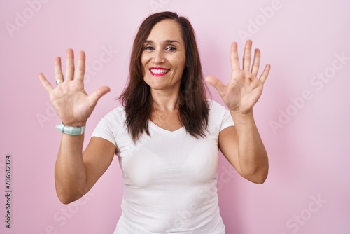 Middle age brunette woman standing over pink background showing and pointing up with fingers number ten while smiling confident and happy.