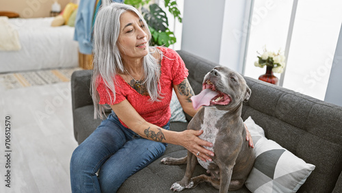 Joyful middle age woman sitting on the sofa at home, playing with her beloved grey-haired dog, laughing and expressing pure happiness