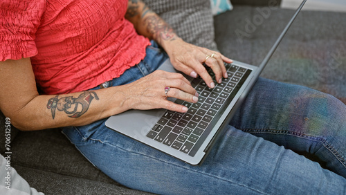 At home, a hardworking woman's hands click away on her laptop, comfortably ensconced on her cozy sofa amidst the familiar backdrop of her living room, fully utilising technology to work online.